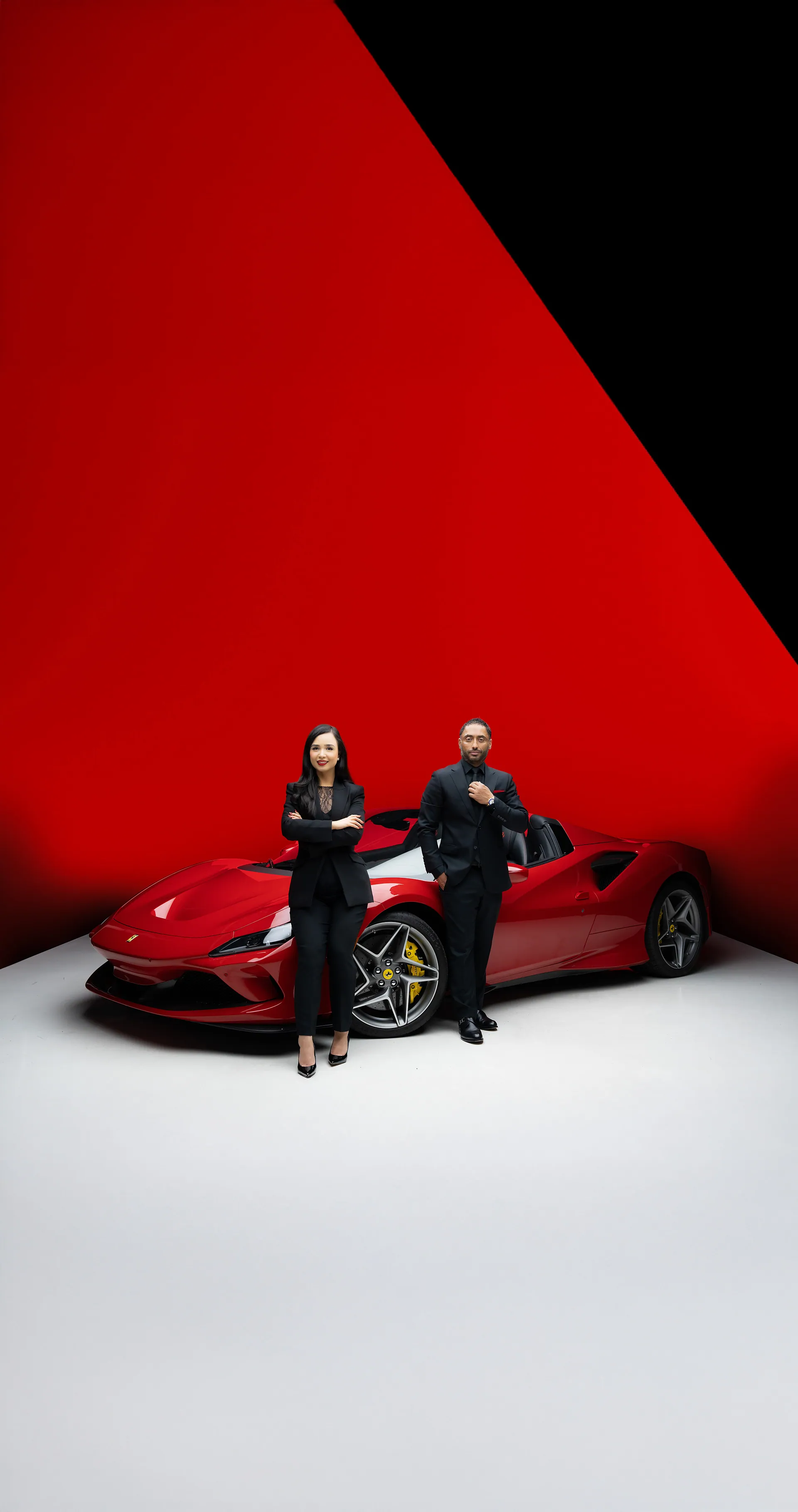 Man and woman in black outfits standing beside a red sports car against a red and white studio backdrop